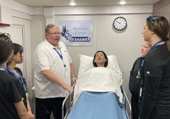 A group of nursing students and an instructor stand around a hospital bed with a patient simulator. The instructor, wearing a white polo and a blue lanyard, is speaking to the students, who are dressed in black scrubs and matching blue lanyards. A sign on the wall behind them reads “Students First” and displays the Keiser University Seahawks logo. A white clock and medical equipment are visible on the wall above the bed.